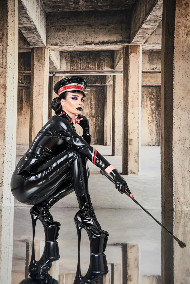 Kara in schwarzem Glanzoutfit mit Schirmmütze und Gerte zwischen Betonstützen.